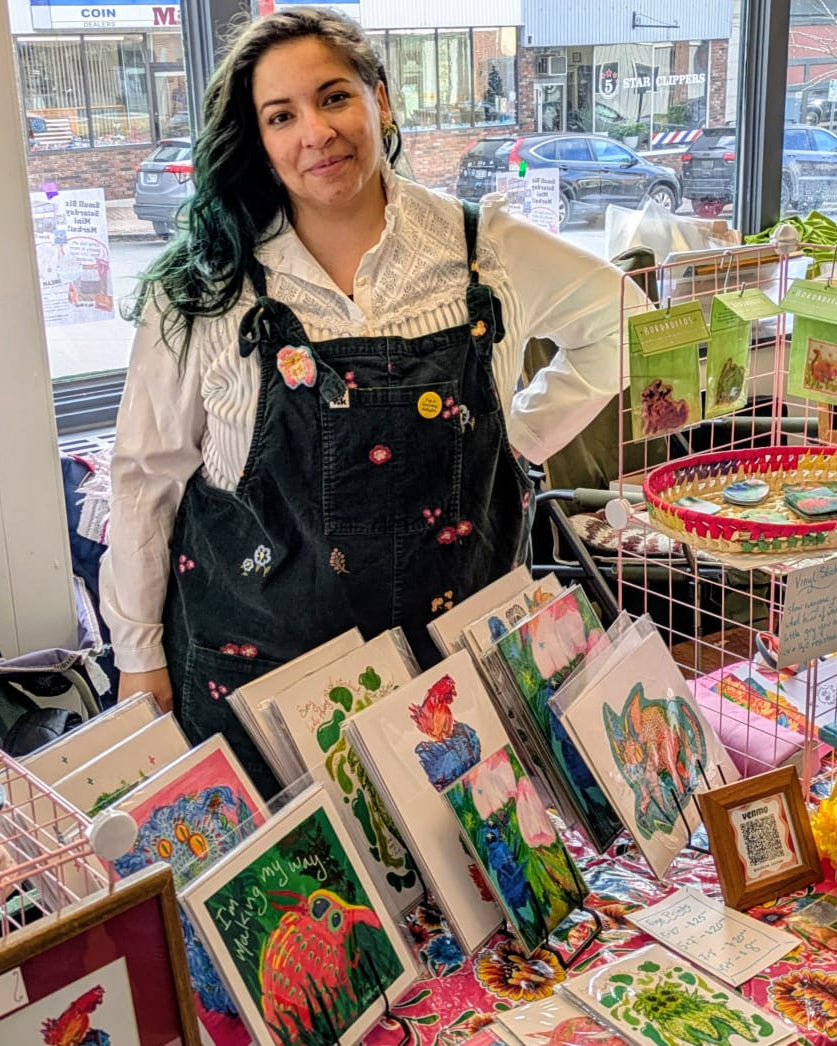 Woman standing behind a table displaying colorful art pieces in an indoor setting. 