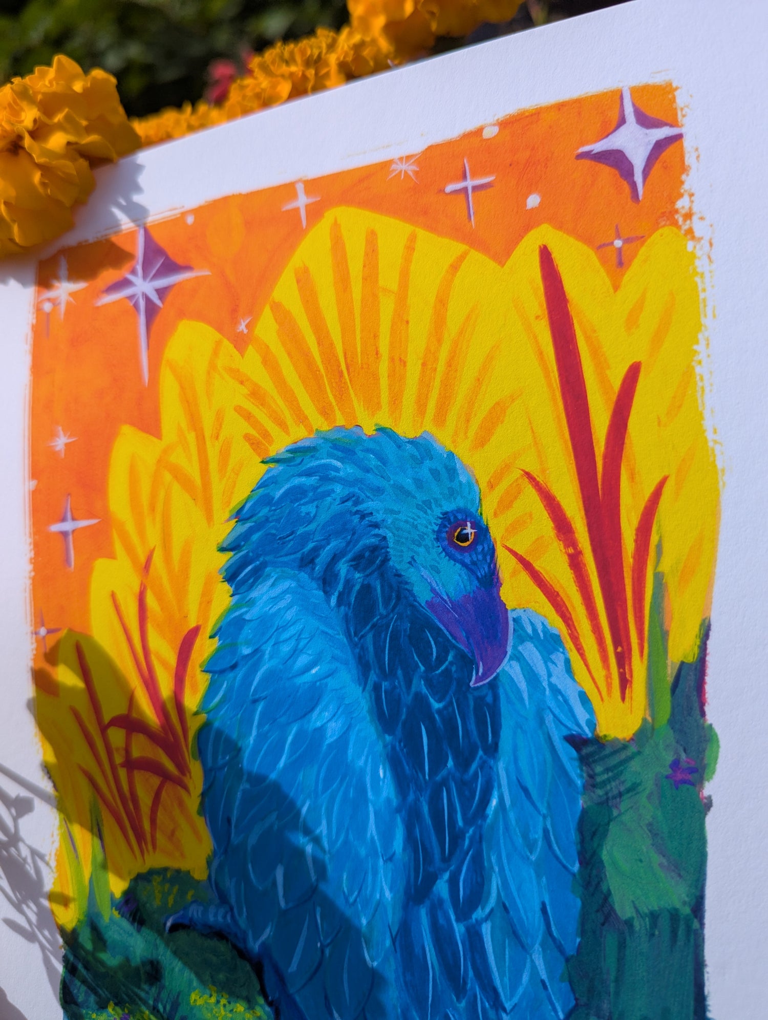A close-up photo of an art print feature "Scavenger." The print is resting on marigolds in a garden, and the photograph is focused on the bright gold eyes of the blue vulture set against a regal orange backdrop. 